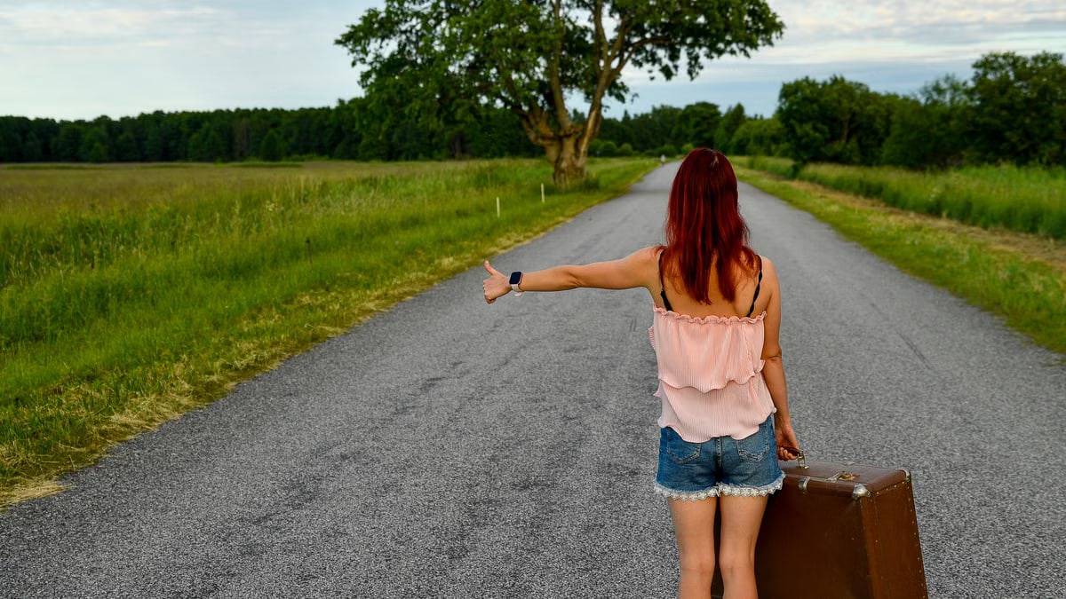 Woman hitchhiking on a rural road in Vihasoo, Estonia, carrying vintage luggage.